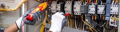 Sähköalan työntekijä tekee mittausta mittalaitteella, työntekijästä näkyy kuvassa ainoastaan kädet.