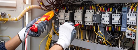Sähköalan työntekijä on tekemässä mittausta mittalaitteella. Työntekijästä näkyy kuvassa ainoastaan kädet.