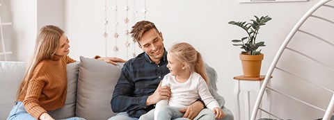 Perhe istuu sohvalla ja hymyilee toisilleen. Lapsi istuu toisen vanhemman sylissä. 