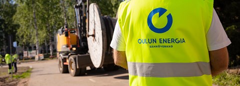 An employee of Oulu Energia Sähköverkko Oy, standing with his back to the camera, looks at the excavator in front of him on the road.