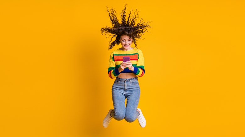 Nuori kiharatukkainen nainen hyppää oranssia taustaa varten kännykkä kädessään